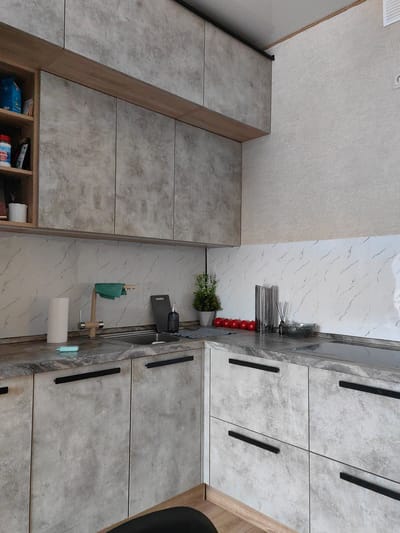 A modern kitchen with grey, textured cabinets and a marble-patterned backsplash is depicted. The scene is well-lit, suggesting daytime. On the countertop, a roll of paper towels, a blue dish sponge, a faucet, and a kitchen sink are visible. To the right, a cutting board, a decorative soap dispenser, a potted plant, and a row of red tomatoes are arranged. Further along the counter, a glass vase and a bottle of reed diffuser sit near a stovetop. The cabinets have sleek black handles. On the left side of the image, a wooden open shelving unit holds various items, including blue packaging and a white container. The overall impression is a clean and organized kitchen space. No people are present in the image. The location is identified as Aprelevka, Russia, but this context is not directly evident from the visual elements alone.