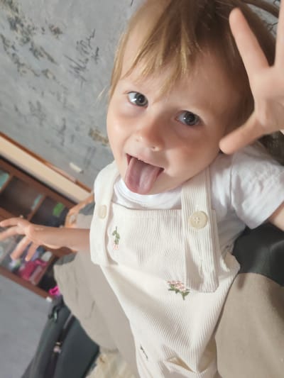 The image shows a close-up, Dutch angle shot of a young child with blonde hair sticking their tongue out. The child is wearing a white t-shirt and off-white corduroy overalls with embroidered pink flowers and green leaves. The child's left hand is raised, appearing to be reaching towards the camera, and their right hand is also visible, partially obscured. The background consists of a textured, grey wall with some indistinct markings, and a portion of a wooden piece of furniture is visible on the left. The scene appears to be indoors, with a soft, natural light source. No specific location in Mezhdurechensk, Russia can be determined from the image.