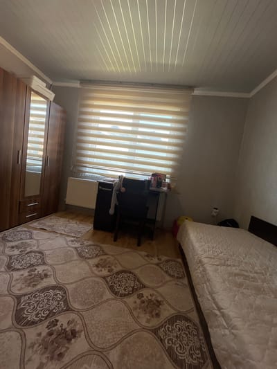 The interior shot reveals a bedroom setting, likely in a residential building. The focal point includes a bed with a textured white cover, positioned next to a beige rug with floral patterns. Adjacent to the window, which is covered by vertical blinds allowing natural light, is a desk with shelves holding decorative items. A white radiator is also visible. In the lower portion of the frame, there is a wooden wardrobe with mirrored doors. The room's walls are light gray, and the overall scene suggests a simple and lived-in environment. The location in Slivo Pole, Bulgaria, is not apparent from the image.