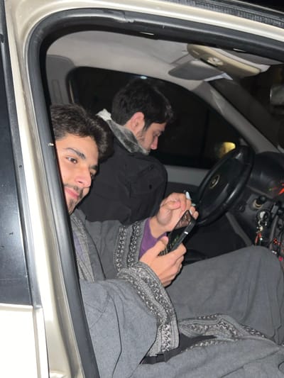 Two men are inside a car, seen through an open window. One lies with his head near the sill, gazing toward the viewer. The second man is further back, holding a phone and what appears to be a cigarette. He's wearing a gray jacket with a patterned trim, and a purple sleeve is visible underneath. The car's interior is dark, with glimpses of the steering wheel and dashboard.