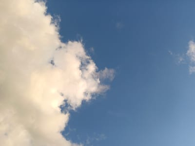 The image displays a clear blue sky with scattered clouds. On the left side of the frame, a large, dense cloud formation dominates, appearing bright and illuminated, possibly by sunlight. These clouds are predominantly white with hints of yellow and grey, suggesting depth and texture. The sky transitions to a deeper blue towards the right, with fewer and more dispersed cloud wisps. A small, dark speck, likely a bird, is visible in the upper right quadrant of the sky. The image appears to be taken from an outdoor perspective, looking upwards. The scene does not contain any elements that would specifically identify it as being within the city of Baranavichy, Belarus.
