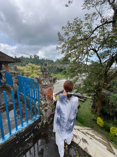 A woman with reddish-brown hair pulled into a ponytail is seen from behind, standing on a wet, stone stairway. She is wearing a white, patterned poncho and white pants. To her left, a vibrant blue metal fence lines a pathway. Beyond the fence, there are traditional Balinese structures with terracotta tile roofs and stone pillars adorned with moss. A river flows in the midground, spanned by a light-colored truss bridge in the distance. Lush green trees and vegetation surround the area, with a large tree dominating the right side of the frame. The sky is overcast with gray clouds. The scene appears to be the Goa Gajah elephant cave complex in Klungkung, Indonesia, a popular tourist attraction known for its historical significance and natural beauty.