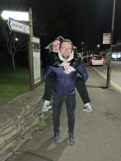 At night in Windermere, United Kingdom, a man in a blue puffer jacket, dark jeans, and blue and red sneakers gives a woman a piggyback ride. The woman wears black pants, white and silver sneakers, a black leather jacket, and glasses. They are near a signpost that indicates directions to the "Station" and "To Lake." The signpost also contains information about the area. The background shows a road with a car driving on it, streetlights, and buildings. The ground is a mix of concrete and a stone wall with grass and greenery.