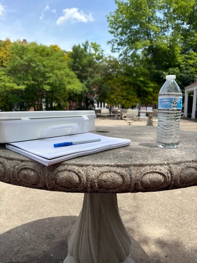 A water bottle sits on a round concrete table next to a white clipboard with papers and a blue pen on top. The table is located outdoors, with green trees and a light blue sky visible in the background. The OCR text on the water bottle includes "DISTRIBUTED BY: Wal-Mart Stores, Inc., AR 72716 PURIFIED BY: Reverse Osmosis PLASTIC BOTTLE". The location cannot be determined as being in Edison, New Jersey as there are no other clues for the location to be identified.