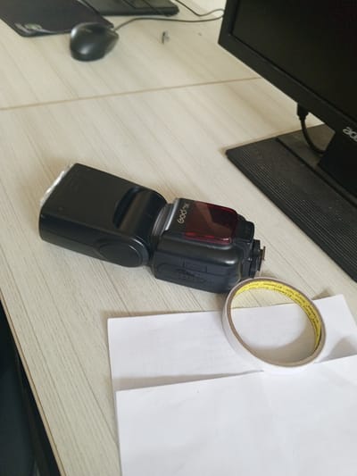 The image depicts a desk with a camera flash, a roll of double-sided tape, and a computer monitor. The camera flash is black and has "Godox" branding. The roll of tape is yellow and white, with text that is difficult to read. The computer monitor is black and appears to be an Acer brand. The desk surface is light-colored with a wood grain texture. There is also a black computer mouse visible in the upper left portion of the image. The scene is indoors and appears to be an office or workspace. There is no indication of the city of Chutula, Mozambique, in the image.