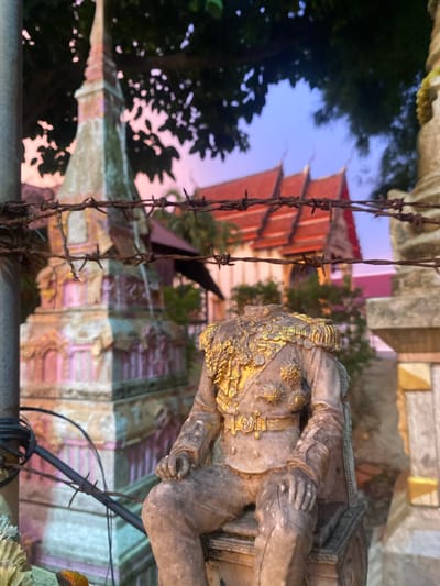 A headless statue of a person in a formal military-style uniform, adorned with gold embellishments, sits in the foreground. The statue is weathered and appears to be made of stone or concrete. Behind the statue, a barbed wire fence stretches across the frame, obscuring the background. In the middle ground, there is a tall, ornate structure with a spire, possibly a monument or a part of a temple, painted in pastel pink and green. Further in the background, the distinctive red-tiled roofs of a temple are visible against a twilight sky that transitions from purple to blue. The setting is likely in Tambon Choeng Thale, Thailand, given the architectural style of the temple and the tropical foliage suggested by the trees and leaves. The overall scene evokes a sense of decay and historical significance, with the juxtaposition of the weathered statue and ornate structures.