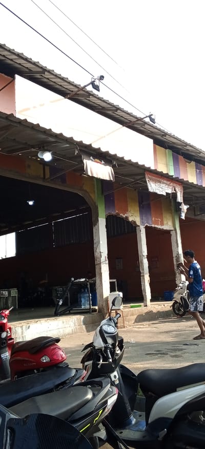 This is a full-frame, eye-level, outdoor shot taken at a car wash or repair shop in Kiangroke, Indonesia. The scene is brightly lit by natural sunlight. In the foreground, three scooters are parked in a line. The leftmost scooter is red, followed by a black and white scooter, and then a white and black scooter. The scooters are positioned facing left and right, with their seats visible.

In the midground, a person wearing a blue t-shirt and patterned shorts is standing near a white scooter, looking down at a mobile device. They appear to be holding the device with both hands. Behind this person is an open-air structure with a corrugated metal roof supported by concrete pillars. The walls of the structure are painted with vertical stripes of orange, yellow, purple, and green. Inside the structure, there are large blue water jugs on a shelf, and a few dark-colored vehicles. A ceiling light is visible inside the structure.

In the background, the sky is overcast and bright white. The overall environment suggests a commercial area, likely a service station given the presence of multiple vehicles and the car wash equipment. The architecture and the presence of scooters are indicative of an Indonesian setting.