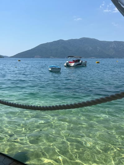 The image shows a view of the Bay of Kotor in Risan, Montenegro, on a clear, sunny day. The foreground is dominated by a thick, dark rope stretched horizontally across the bottom of the frame, obscuring part of the very clear, shallow water below. The water itself is a vibrant turquoise and green, with sunlight illuminating the sandy bottom and a school of small fish swimming in the shallows.

In the middle ground, two boats are visible on the calm, blue water. To the left, a small, light blue and white dinghy is tied to a yellow buoy. Further to the right, a white motorboat with a red interior and a canopy is anchored, also near a yellow buoy. More yellow buoys are scattered across the bay, indicating marked areas.

In the background, a large, lush green mountain range rises steeply from the water's edge, its slopes covered in dense vegetation. The base of the mountains is dotted with small buildings, suggesting a coastal settlement. The sky above is a clear, brilliant blue with a few wispy white clouds near the top right corner. The overall impression is one of tranquility and natural beauty.