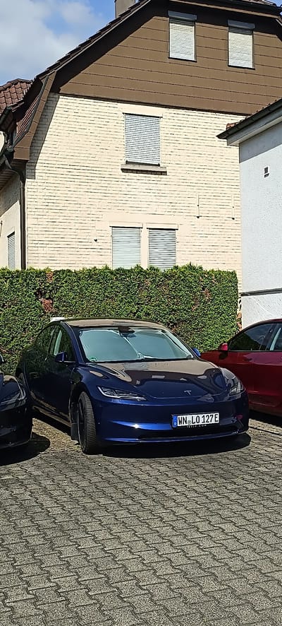 In Endersbach, Germany, a blue Tesla Model 3 is parked on a paver driveway in front of a light-colored brick house with dark brown trim and a dark roof. The house has several windows with rolled-down shutters. A trimmed green hedge separates the driveway from the house. A portion of a dark-colored vehicle is visible to the left of the Tesla, and a dark red Tesla is parked to the right, partially obscured by the blue Tesla. The sky is partly cloudy.