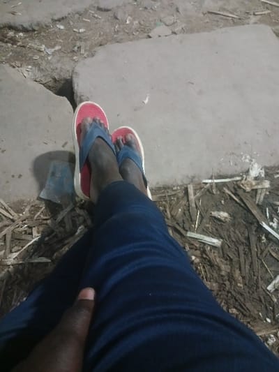 A person wearing red and blue flip-flops stands on a concrete ledge. To the right, the person wears dark blue pants. Debris and small sticks litter the ground. A shadow falls across the ledge near the feet.