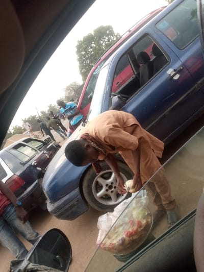 An image, captured from the interior of a vehicle looking out the passenger window at an upward angle, depicts a busy roadside scene in Tataura, Nigeria. In the immediate foreground, a clear plastic bag containing salad ingredients (including visible tomatoes, lettuce, and onions) rests within a metallic bowl, positioned on a surface outside the vehicle. A dark blue four-door vehicle, possibly a sedan or hatchback with a red roofline accent, occupies the central mid-ground. Its front passenger door is open, exposing the interior. A young male, dressed in a light brown tunic and pants, with a multi-colored scarf around his neck, is bent over near the front passenger wheel of the blue vehicle, actively handling a pale yellow object in his hands. To the left, a dark-colored sedan is partially visible behind the blue vehicle. In the background, several individuals are present on an unpaved dirt road, including one male in a blue and white striped shirt leaning against the blue vehicle. Trees and distant foliage are visible under a bright, overcast sky.