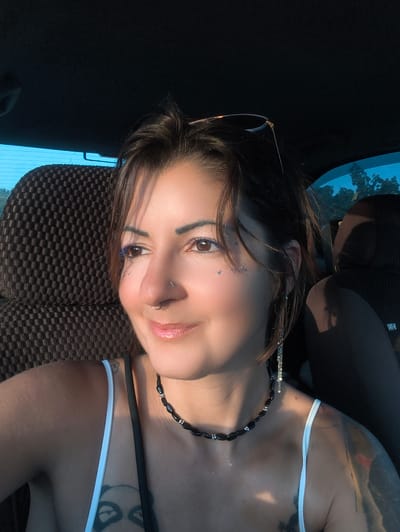 An adult female is depicted seated inside a vehicle in the Mate Yehuda Regional Council, Israel. She possesses dark brown hair, with strands framing her face, and sunglasses are positioned on top of her head. Her face is illuminated by strong, warm, natural light from the right side of the frame, creating highlights on her skin. She is smiling faintly and looking towards her left. Her brown eyes feature blue eyeliner on the upper lash line, and small, reflective, glitter-like particles are present beneath her left eye. She has a circular septum piercing and a stud piercing on her left nostril. Her lips are slightly parted and appear glossy. She is wearing a white spaghetti strap top, and a black beaded necklace with silver accents is around her neck. A black strap is visible crossing her upper chest, possibly from a bag or an undergarment. Tattoos are visible on both shoulders: a partial panda face design on her right shoulder/chest area and an abstract design on her left shoulder. A long, dangling earring with multiple silver elements hangs from her left ear. The car's interior features a dark brown, textured, checkered seat behind her. Through a tinted window on her left, a bright blue sky and green foliage (trees) are visible. The car's ceiling is dark.