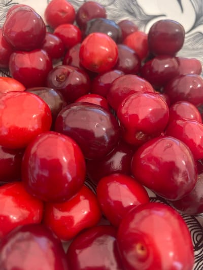 The image displays a close-up, high-angle view of numerous ripe cherries, predominantly red and dark red varieties, piled together in what appears to be a bowl. The cherries exhibit a glossy, reflective skin texture, indicating freshness. Bright red cherries are interspersed with deeper, almost black-red varieties, suggesting a mix or varying stages of ripeness. Some cherries show subtle indentations where stems would have been attached. The background consists of a partial view of a white surface with a black, wavy, and abstract linear pattern, likely the interior or rim of the container holding the cherries. No individuals or specific actions are visible. The environment is limited to the immediate context of the cherries and their container, offering no discernible geographical markers or indications of a specific city such as Kipilovo, Bulgaria.