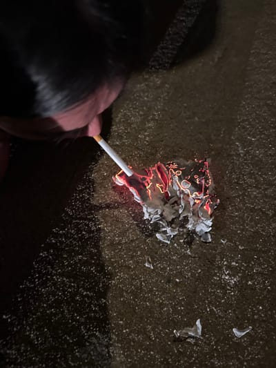 The scene is set in Roanne, France, depicting a close-up, dimly lit, high-angle view of an individual lighting a cigarette. The upper left quadrant of the frame shows the lower face, chin, and dark hair of a person. A white cigarette with a light brown filter is held in the individual's mouth or between fingers, extending horizontally towards a burning object on the ground. This object is a piece of paper, partially consumed by fire, exhibiting vibrant orange-red embers and flames, surrounded by curled fragments of white ash and charred material. The burning paper rests on a dark, granular ground surface, consistent with asphalt or coarse concrete, which is speckled with numerous small, light-colored particles. The tip of the cigarette is in direct contact or very close proximity to the active flames and embers of the burning paper, indicating the act of ignition. The primary illumination for the scene originates from the burning paper itself, casting a warm glow on the immediate area against the overall dim environment.