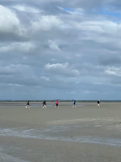A group of five individuals walk across a wide, flat, wet sand expanse under a cloudy sky. The people are spaced out, walking from left to right across the frame. They are all wearing casual clothing, with some carrying backpacks. The sand shows ripples and shallow pools of water, indicating it is likely a tidal flat. In the distance, a faint treeline and possibly buildings are visible on the horizon. This scene is consistent with the appearance of the bay surrounding Mont-Saint-Michel, France, a famous tidal island and commune.
