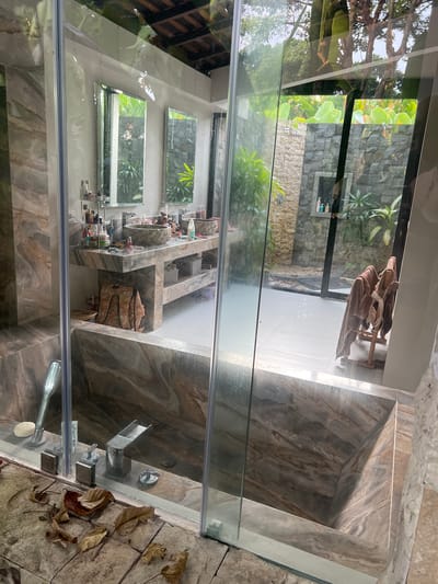 The image depicts the interior of a bathroom in Kerobokan, Indonesia, as viewed through a glass shower partition. The bathroom features marble-like tiling. Through the glass, one can see a reflection of a vanity area with multiple shelves holding various toiletries. Windows above the vanity reflect an exterior view of lush greenery. Beyond the shower, an outdoor space is visible, containing plants, a stone wall, and a clothes rack with clothing. The surrounding area appears to be a garden. The flooring outside the shower area is stone-tiled, with some fallen leaves scattered on the surface. The shower fixture, including the faucet and showerhead, are visible.