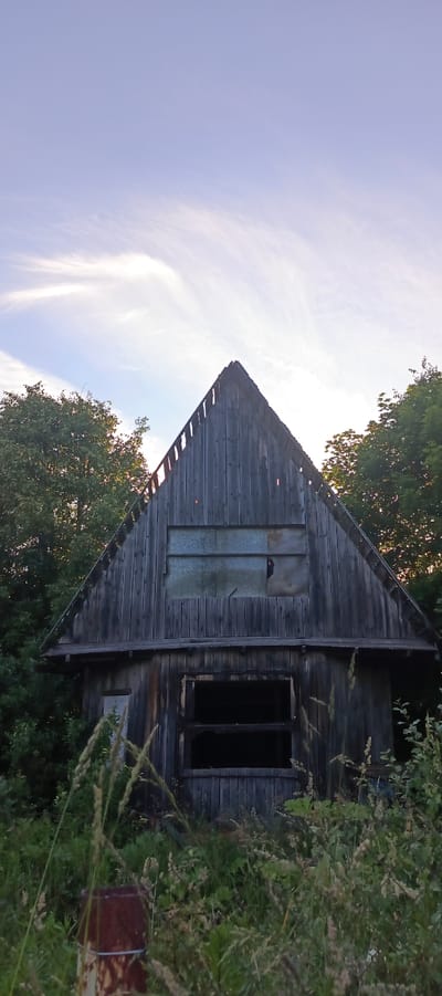 An abandoned wooden building with a gabled roof stands amidst overgrown vegetation. The building's facade is weathered and features two dark, gaping window openings on the lower level and a boarded-up window with a translucent material on the upper level. Tall grass and foliage obscure the base of the structure. The sky above is a pale blue with wispy clouds. The scene appears to be located in Merchalovo, Russia.