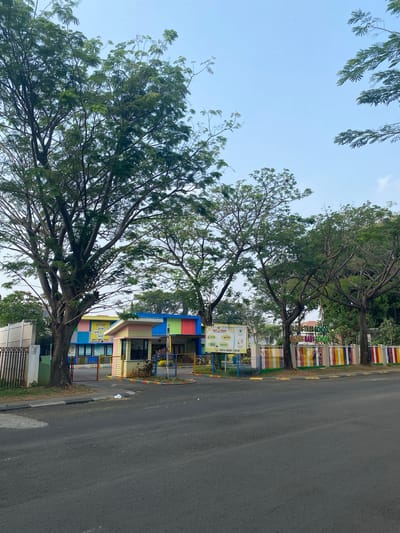 The image depicts the exterior of a brightly colored school building, likely a kindergarten or preschool, situated in East Jakarta, Indonesia. The building features a vibrant facade with sections painted in yellow, pink, blue, and green. A security booth is positioned at the entrance, which is flanked by a colorful fence adorned with red, yellow, blue, and white vertical bars. Large trees with lush green foliage are scattered throughout the scene, providing shade and framing the school. A paved road runs in front of the school, with a sidewalk separating it from the property. The sky above is a clear, pale blue. There are no people visible in the image. The overall impression is that of a well-maintained educational facility set against a backdrop of natural greenery.