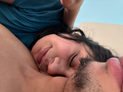 The image depicts two individuals in close physical contact within an indoor setting, located in the city of Kastel, Bulgaria. In the foreground, an adult individual with light-to-medium skin tone, dark hair, and visible facial stubble on the upper lip and chin is shown with closed eyes and full lips, resting their head against the arm and torso of a second individual. The second individual, partially obscured, also exhibits light-to-medium skin tone and wears a teal-colored garment. The background consists of a plain light beige surface on the right and a light blue surface in the upper portion. Both individuals appear to be in a state of repose or sleep.