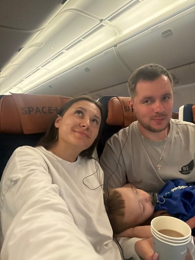A family of three is on an airplane. A man in a gray shirt and a silver cross necklace looks at the camera. Beside him, a sleeping child with a blue hoodie lies on a woman's shoulder. The woman wears a white long-sleeved shirt and gazes upward. Airplane windows are visible on the left. Seat headrests labeled "SPACE+" are visible behind the couple. The man holds disposable cups.