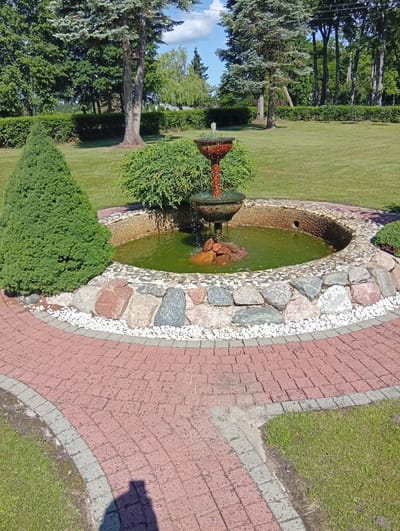 The image shows a garden area with a fountain as the central feature. The fountain is made of a reddish-brown material and has water flowing from its top tier. The fountain is situated in a circular pond with a stone border. Surrounding the pond is a pathway made of red brick pavers, bordered by smaller grey pavers and white gravel. A green, cone-shaped shrub is planted to the left of the fountain, and a larger, bushy green shrub is behind it. The background consists of a neatly manicured green lawn, several mature trees, and a line of trimmed hedges. The sky is visible above the trees, with some white clouds against a blue background. The scene appears to be an outdoor garden in a well-maintained residential or public space. The image is taken from a slightly elevated angle, looking down towards the fountain and pathway. The time of day appears to be daytime, with clear sunlight casting shadows, including a distinct shadow of a person in the foreground on the pathway. The location is stated to be Dirvonėnai, Lithuania.
