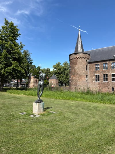 This media file depicts an outdoor scene featuring a bronze statue of a nude figure in a grassy area, with a historic brick building and a conical tower in the background. The statue is positioned on a stone pedestal, and appears to be a classical or artistic representation of a human form. The building behind the statue is constructed of red brick with multiple windows and a dark-colored roof. A tall, pointed tower, also with a dark roof, is visible to the left of the main building.
The setting is a well-maintained lawn with tall grass bordering the paved area around the statue. Lush green trees are present in the mid-ground and background, suggesting a park or garden environment. The sky is a vibrant blue with a few wispy white clouds and contrails from an aircraft. The lighting indicates it is daytime, likely mid-morning or afternoon, with bright sunshine casting shadows.
The overall mood is serene and picturesque, with the juxtaposition of the ancient architecture, the classical statue, and the natural landscape. There is no discernible activity taking place, as the scene appears static and posed. No text is visible within the image. The location context suggests this scene is in Helmond, Netherlands.