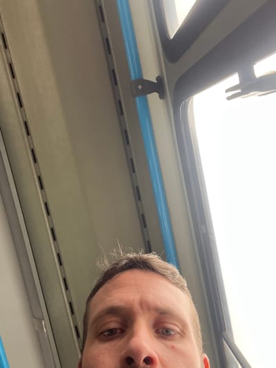 An unusually angled shot captures the partial face of a man in the upper right quadrant, seemingly taken as a selfie or from an upward perspective within a vehicle. His eyes are visible, looking slightly upwards, and he has light brown or greying hair.
The setting is the interior of what appears to be a public transport vehicle, likely a train or bus, situated in Cordoba, Argentina. The ceiling and upper sections of the vehicle are prominently featured. A bright, almost overexposed, white light fills the upper left portion of the image, indicating a large window or skylight above, suggesting it is daytime with bright weather outside.
Below the bright window area, there's a dark grey trim, followed by a lighter grey or beige panel that forms part of the vehicle's interior. A distinctive bright blue horizontal bar runs across the mid-section of the image, potentially serving as a handrail. Below this blue bar, more light grey/beige paneling with horizontal rows of small black slots or vents is visible, suggesting overhead storage or ventilation. A black latch or handle mechanism is visible attached to the ceiling structure, near the window. No discernible text is visible anywhere in the image.
