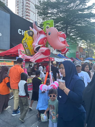 The image captures a vibrant street scene, likely in Makassar, Indonesia, during an outdoor market or festival. A prominent display of colorful balloons, including a yellow car, a pink elephant, and other cartoon-shaped inflatables, dominates the foreground, held by unseen individuals. Several people are visible, dressed in casual attire, many wearing headscarves, suggesting a predominantly Muslim population.

In the mid-ground, a woman in a dark blue hijab sips from a cup while a young child wearing pink sunglasses stands beside her, looking towards the camera. Other individuals, including a man in an orange shirt and children, are also present, suggesting a family outing or community gathering. Tents and stalls, some with red awnings, indicate the presence of vendors. In the background, a tall, modern building rises above the scene, contrasting with the more traditional market setup. The overall atmosphere appears lively and bustling.