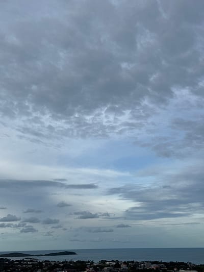 An aerial, landscape photograph captures a cloudy sky over the coastal town of Ko Samui District, Thailand. The sky is filled with a variety of gray and white clouds, with a small patch of lighter blue sky visible towards the center. Below the sky, the vast, calm ocean stretches to the horizon. Along the coastline, a dense collection of buildings, ranging from single-story homes to multi-story structures, are visible, interspersed with dark green trees. Two small, hilly islands are situated in the ocean, adding to the scenic view of this coastal area.
