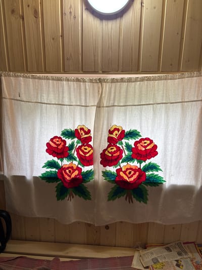 The image shows a close-up of a white curtain with embroidered red and yellow roses and green leaves. The curtain is hung in front of a wooden wall. A light fixture is visible at the top center of the image, casting a warm glow. In the lower part of the image, a wooden shelf is visible, with some newspapers and a red and white checkered cloth on it. The scene appears to be in a rustic indoor setting. There are no people visible in the image. The city of Ильпанур, Russia is not depicted in this image.