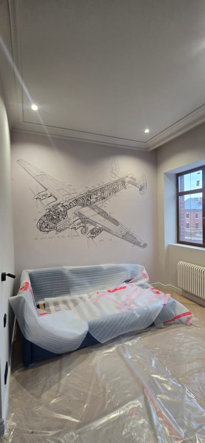The image depicts an interior scene, likely a living room, in a property located in Русская Деревня-2 Кп, Russia. Dominating the wall is a large black and white schematic drawing of an airplane. In the foreground, a navy blue sofa is partially covered in protective foam packaging, with red tape visible. The floor is covered with clear plastic sheeting, suggesting ongoing renovations or that the furniture is new and being protected. To the right, a window with a wooden frame looks out onto a building with red brick elements. A white radiator is situated beneath the window. The ceiling is white with recessed lighting fixtures and decorative molding. Two black wall-mounted light fixtures are visible on the left side of the image.