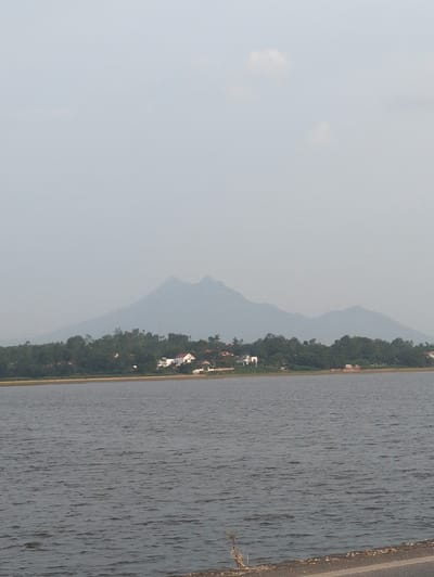 The image depicts a wide, calm body of water, likely a lake or a river, with a shoreline in the mid-ground. Along the shoreline, there is a stretch of vegetation, predominantly trees, dotted with several buildings. These buildings appear to be houses or small structures, some with white walls and red roofs, suggesting a rural or semi-urban settlement. In the background, hazy, blue-tinged mountains rise against a pale sky with a few scattered clouds. The overall atmosphere is serene and somewhat muted due to atmospheric haze. This scene is likely located in or near Dao Xa, Vietnam, given the presence of mountains, water, and vegetation characteristic of the region.