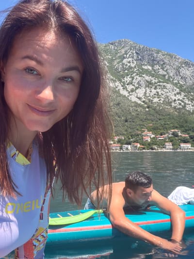 A selfie is taken on the water, with the partial face of a woman with dark brown hair, fair skin, and green eyes dominating the foreground. She wears a light-colored shirt with the word "O'Neill" printed on it. Behind her, a man with dark hair is seen half-submerged in the water, holding onto a teal and white inflatable paddleboard. In the background, a mountain range covered with green vegetation rises steeply from the water's edge. Buildings are visible along the shoreline, suggesting a populated area. The clear blue sky indicates a sunny day. The location is Dražin Vrt, Montenegro, as indicated by the recognizable landscape.
