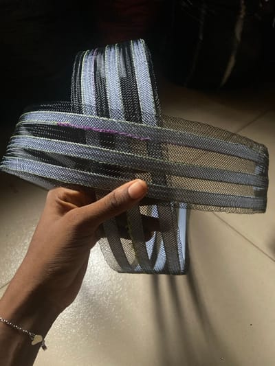 A hand with dark skin holds a crafted object of stacked, striped mesh. The mesh is black with metallic silver and gold lines. The object's shape suggests a container or headpiece, constructed from layered loops of the mesh. The hand has a simple silver bracelet with small heart charms. The background shows a shiny black surface and a lighter, tile-like surface.