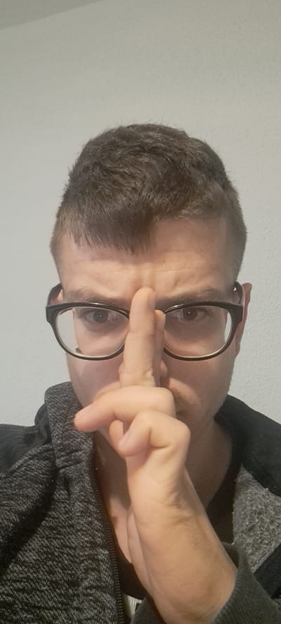 A man is posing for a selfie. He is wearing glasses and a grey zip-up sweater. His right index finger is touching the bridge of his glasses. The background is a plain white wall. There is no indication of Mineralni Bani, Bulgaria in the image.