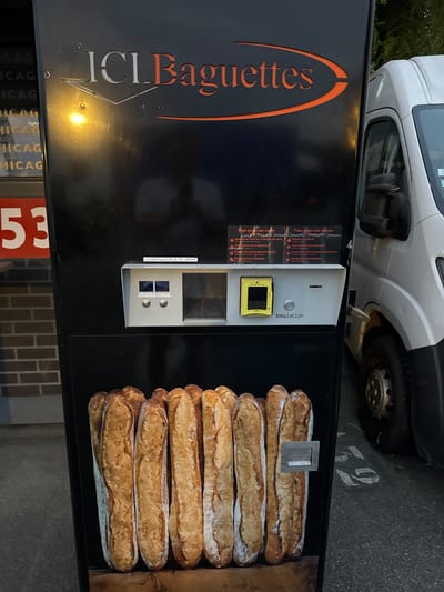 This image captures a baguette vending machine labeled "ICI Baguettes" located outside in Grenay, France. The vending machine is primarily black with orange and white lettering. It features a slot for payment, a keypad, and a yellow card reader. Below the payment interface, a display shows a close-up image of several baguettes. To the right of the vending machine, the front of a white van is visible, parked adjacent to the machine. The background includes a brick wall and some greenery, suggesting an urban or semi-urban outdoor setting. The ground beneath the machine is asphalt, with white markings partially visible, indicating a parking area or public space.