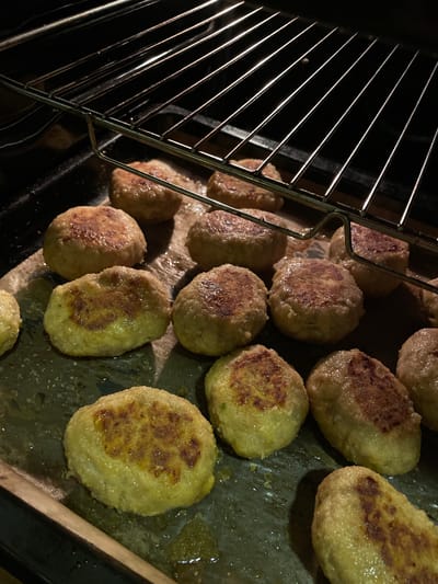 A batch of golden-brown, oval-shaped patties are arranged on a baking sheet inside a dark oven. The patties appear to be cooked or baking, with some showing slightly darker, crisped edges. The oven rack is visible above the patties, reflecting the interior light. The baking sheet is dark and shows some grease or cooking residue. The overall lighting is dim, with light primarily coming from the oven's interior, creating shadows and highlighting the texture of the patties. The scene depicts food being prepared in an oven. The location in La Línea de la Concepción, Spain, is not discernible from the image itself.