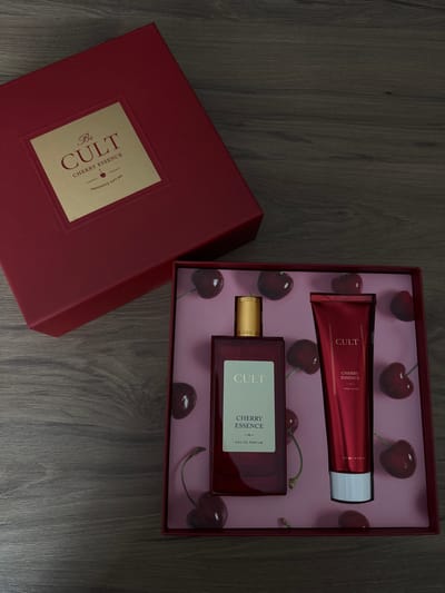 A red gift box for a fragrance set is open, revealing a perfume bottle and a tube of hand cream. The interior of the box is decorated with a pink background featuring a pattern of cherries. The perfume bottle is dark red glass with a gold cap and a label that reads "CULT CHERRY ESSENCE EAU DE PARFUM". The hand cream tube is also red and has a label that reads "CULT CHERRY ESSENCE HAND CREAM". The gift box itself is dark red and has a gold label on the lid with the text "Be CULT CHERRY ESSENCE FRAGRANCE GIFT SET". The items are arranged on a dark wooden surface. There are no people or animals present in the image. The location in the city of Murom, Russia is not discernible from the image.