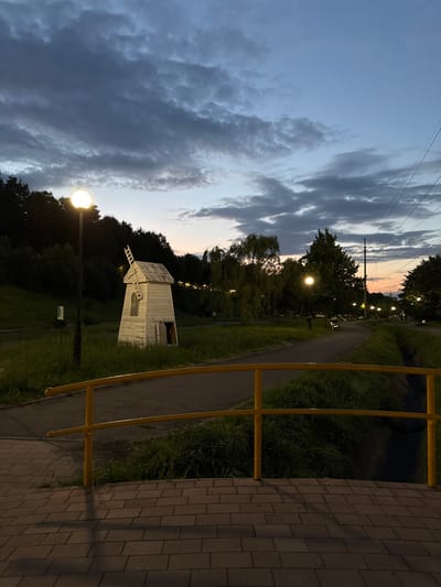The image depicts a park-like setting at dusk, with a pathway winding through grassy areas and trees. A small, white, rustic windmill structure stands on the left side of the pathway. Several streetlights are visible, casting a warm glow on the surroundings. A yellow metal railing runs along the right side of the pathway, adjacent to a brick-paved area. The sky is filled with dramatic, dark clouds, suggesting an overcast or transitional weather condition. The overall mood is peaceful and serene, with the soft lighting contributing to a tranquil atmosphere. The scene appears to be in a public park or recreational area, possibly in Vladikavkaz, Russia, as indicated by the location context. No people are visible in the frame. The time of day is clearly evening, as indicated by the dim natural light and illuminated streetlights.