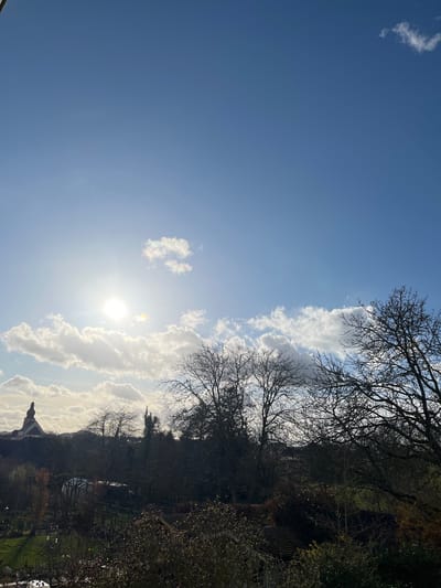 The image displays an outdoor landscape scene situated in Foetz, Luxembourg. The upper two-thirds of the frame feature a clear blue sky with a prominent, intensely bright sun positioned slightly left of center, partially obscured by scattered white cumulus clouds; some clouds exhibit iridescence. The lower third of the image is characterized by a mix of natural and semi-urban elements, primarily silhouetted due to backlighting. On the far left, a church steeple with a conical roof and a cross is visible, rising above distant structures. Below the horizon, a dense collection of bare deciduous trees and some evergreen foliage dominates the mid-ground. A small, transparent-paneled greenhouse structure is discernible amidst the trees. The immediate foreground consists of dark, dense vegetation, including shrubs and the upper portions of trees, partially obscuring the lower ground. No individuals or explicit actions are visible within the frame.