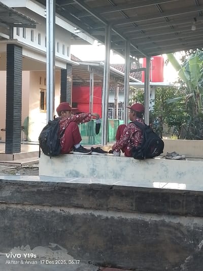 Two children wearing red patterned shirts and matching caps sit side-by-side on a white concrete ledge. Each wears a black backpack. One child holds up a black object, possibly a shoe. Additional pairs of shoes rest on the ledge beside them. The background features buildings, a red water tank, and greenery, partially obscured by a metal structure. The photo was taken in daylight. The bottom of the image contains a watermark.