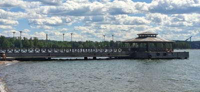 The image depicts a scenic view of a pier extending into calm, rippling water, likely a lake or sea. The pier is constructed of wood with a dark, possibly metal railing featuring a criss-cross pattern. At the end of the pier, a hexagonal structure with a dark roof and open sides serves as a covered shelter or gazebo. Several streetlights with dark, circular lamps are positioned along the pier, suggesting it is illuminated for evening use.

On the pier, two people are visible. One individual, wearing a dark top and shorts, sits with their legs extended. Another person, further down the pier and closer to the water's edge, appears to be either sitting or lying down. The overall scene suggests a leisurely summer day in a location like Lahti, Finland. The sky is filled with dramatic, white cumulus clouds against a bright blue backdrop, indicative of fair weather and possibly mid-afternoon or early evening light. The water is a tranquil blue-green, reflecting the sky and surrounding trees. In the background, a dense line of green trees borders the water, with hints of buildings and a tall industrial chimney visible among them. The camera angle is unusual, as the image is rotated 90 degrees counter-clockwise, making the water appear to be at the top of the frame and the sky at the bottom.