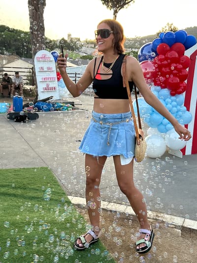 In Sant Vicenç De Montalt, Spain, a woman with medium-length brown hair and sunglasses stands outdoors, holding a black smartphone in her right hand. She is wearing a black cutout sports bra with pink accents, a light-wash denim mini-skirt with a distressed hem, and a decorative gold chain belt with star charms. She has a small woven crossbody bag with a brown leather strap. Her left arm is extended slightly to her side, and she is wearing platform sandals with pastel-colored straps. Numerous bubbles float around her, obscuring parts of the background.
To her left, two men are seated on a bench. The man on the left wears a dark t-shirt and dark shorts, while the man on the right wears a grey t-shirt and khaki shorts. Both are looking at their phones. Behind them, a white shelf unit displays multiple rows of pink cups and red and white striped containers.
In the background, there is a tall tree on the left, and a cluster of red, blue, and white balloons to the right, with white and red striped fabric behind them. The setting appears to be a paved area next to a patch of artificial turf, with a slight incline leading to buildings and a hill in the distance, suggesting a town or village. The lighting indicates it is late afternoon or early evening.