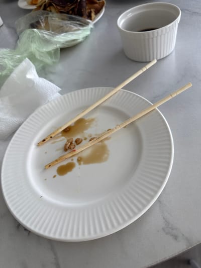 The image depicts a table setting after a meal, specifically on a marble-patterned surface. In the foreground, a white, ribbed plate contains remnants of food, including a few pieces of what appears to be nuts or seeds, along with spilled soy sauce or a similar dark liquid. A pair of wooden chopsticks rests diagonally across the plate, also showing traces of the sauce. To the left of the plate, a crumpled white paper napkin is visible. In the background, a white ramekin filled with a dark liquid, likely soy sauce, is placed on the table. Behind the plate, a portion of a plastic bag filled with what looks like crepes or thin pancakes is partially visible. The overall scene suggests the conclusion of a meal, with the dining area in Нижнеяркеево, Russia. There are no individuals present in the image.