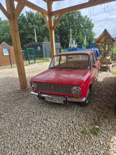 A red Lada car is parked on a gravel lot under a wooden structure with a thatched roof. The car is facing forward and is slightly to the right of the center of the image. The car's license plate reads "B709OP 763 RUS". In the background, there are trees, a fence, and some buildings. The sky is overcast. The scene appears to be in the city of Volzhskii, Russia.