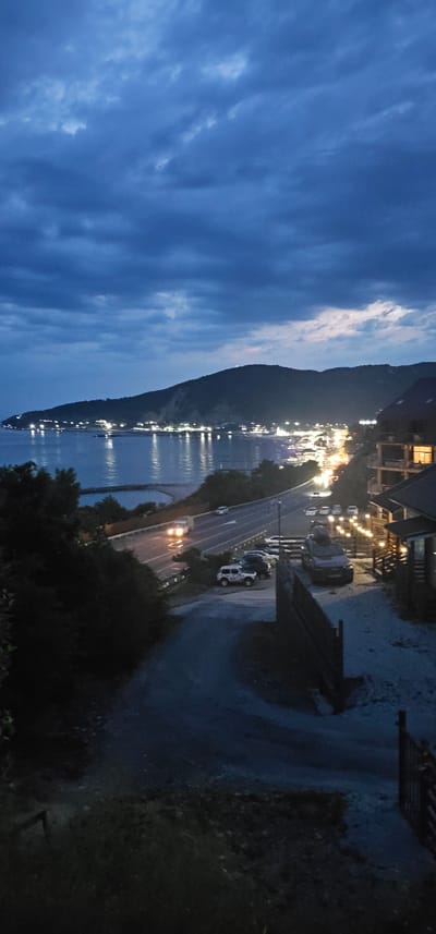 The image depicts a twilight scene in Lermontovo, Russia, overlooking the Black Sea. A coastal road with several vehicles is visible, with lights from a nearby town reflecting on the water. The sky is filled with dark, cloudy formations, indicative of evening or impending weather. The foreground features a pathway leading down towards the road, with trees and foliage on either side. To the right, a building with illuminated windows suggests residential or hospitality structures, possibly hotels or apartments. The overall atmosphere is serene, yet dynamic due to the presence of the road and vehicles.