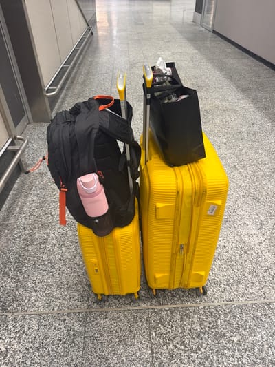 This image depicts two bright yellow suitcases and a black backpack with an attached pink water bottle. The luggage is positioned on a speckled grey tiled floor, characteristic of an airport terminal. A black tote bag is resting on top of one of the suitcases. The scene appears to be inside a modern building, possibly an airport, with metal railings and wall panels visible in the background. The lighting suggests an indoor environment. While the image does not directly show the city of Ferno, Italy, it is likely captured in the vicinity of the Milan Malpensa Airport (MXP), which serves Ferno.