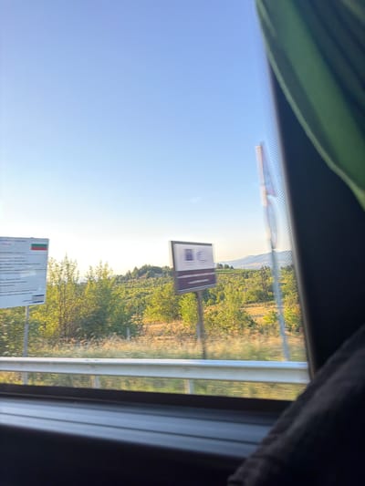The image captures a view from inside a vehicle, looking out a window. The foreground shows a portion of the vehicle's interior, including a dark seat and a reflective window frame. The exterior scene reveals a bright, clear blue sky above a landscape of rolling hills covered in green trees and vegetation. A metal guardrail runs horizontally across the lower portion of the exterior view.
Two signs are visible on the left side of the frame. The leftmost sign is large and rectangular, with text and what appears to be the Bulgarian flag (white, green, and red horizontal stripes). A second, smaller sign is positioned to the right of the first, with a dark background and white icons or text. The setting appears to be a rural area, possibly near a border crossing given the signs. The image is slightly blurred, suggesting motion. The location is identified as Tserovo, Bulgaria.