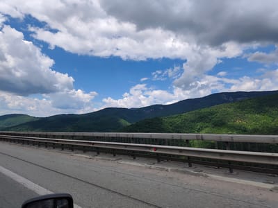 The image shows a view from a vehicle traveling on a bridge in Vrachesh, Bulgaria. In the foreground, the asphalt road is visible, with a white lane marking on the left and a guardrail along the right side of the bridge. The guardrail is made of metal bars and supports. Beyond the bridge, rolling green hills covered in dense forest stretch into the distance. The sky is a vibrant blue, dotted with fluffy white and grey clouds. The overall impression is one of a scenic drive through a mountainous, forested area under a partly cloudy sky.
