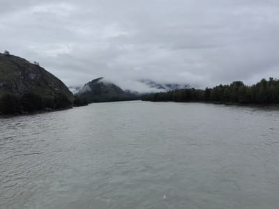 The image depicts a wide, calm river flowing through a mountainous, forested landscape under an overcast sky. On the left, a steep, green, and rocky mountain slopes down towards the river, with a few sparse trees clinging to its crest. On the right, a dense line of dark green trees borders the riverbank. In the distance, more mountains are visible, partially obscured by clouds and mist, creating a hazy, atmospheric effect. The water surface shows gentle ripples. The scene is devoid of any human activity or man-made structures, suggesting a remote, natural setting. The location is identified as Anos, Russia.